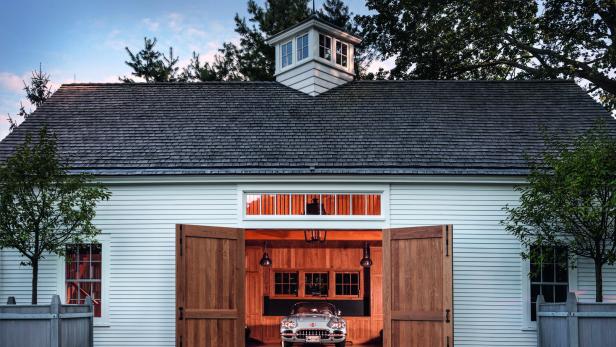 Ein Oldtimer steht in einer offenen, weiß gestrichenen Garage mit Holztoren und gepflegtem Außenbereich.