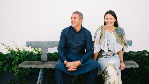 Ein Mann und eine Frau sitzen lachend auf einer alten Holzbank vor einer weißen Wand mit grünen Pflanzen im Hintergrund.