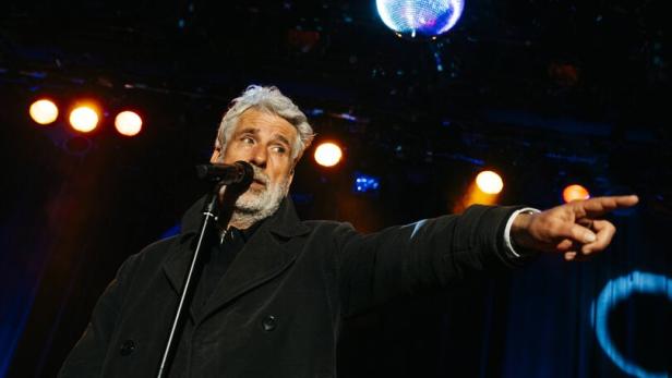 Ein Mann mit grauem Haar spricht in ein Mikrofon und zeigt auf eine Bühne, über ihm hängt eine leuchtende Discokugel.