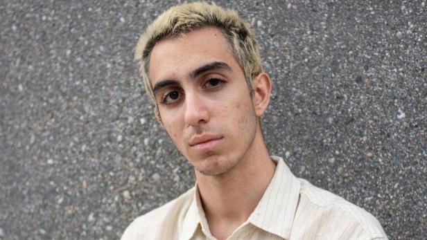 Ein junger Mann mit blond gefärbten Haaren und hellem Hemd steht vor einer grauen Wand.