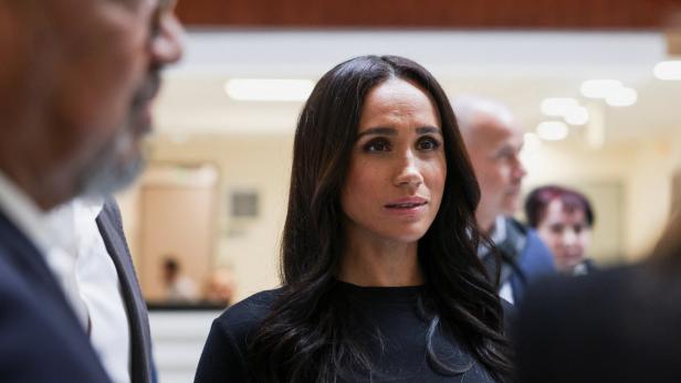 Herzogin Meghan, mit dunklem Haar und schwarzem Oberteil, steht im Gespräch mit anderen Personen.