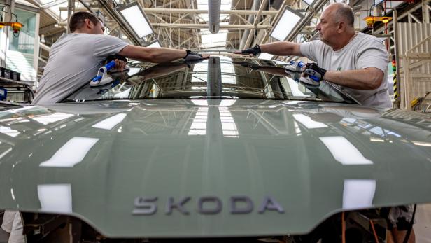 Zwei Arbeiter montieren gemeinsam eine Windschutzscheibe an einem Škoda-Auto in einer Fabrikhalle.