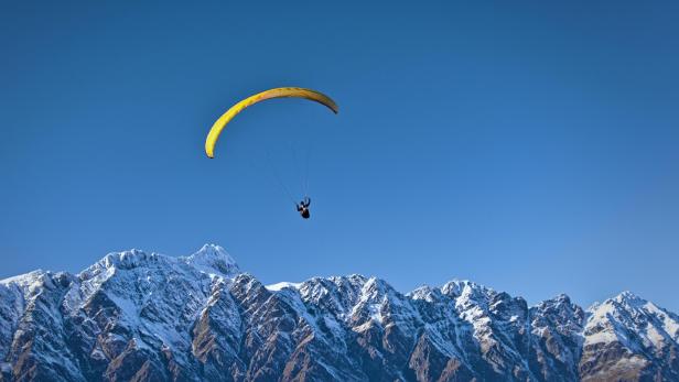 Gleitschirmflieger mit gelbem Schirm über schneebedeckten Bergen und blauem Himmel.