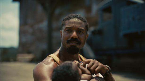 Ein Mann mit ernstem Blick hält ein Baby im Arm, im Hintergrund unscharfe Gebäude und ein Fahrzeug.