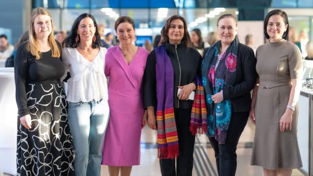 Sechs Frauen stehen nebeneinander und lächeln in einer hellen, modernen Umgebung.