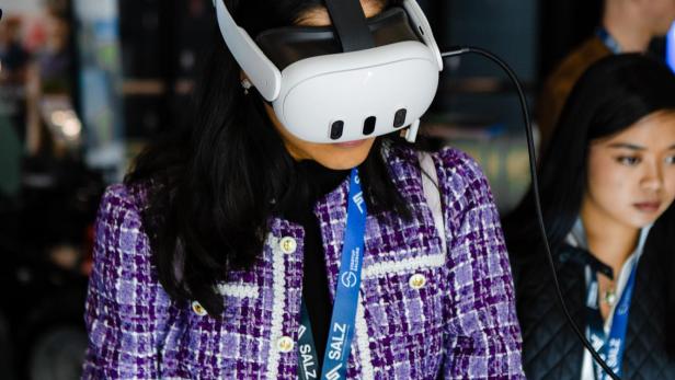 Eine Frau trägt eine weiße VR-Brille, steht vor einem Tisch und schreibt mit einem schwarzen Stift auf die Oberfläche.  Sie trägt einen lila Blazer im Channel-Stil.