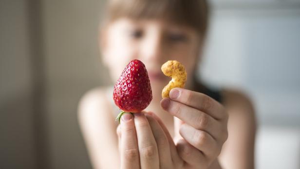 Das Bild zeigt eine unscharfe Aufnahme eines Kindes, davor hält es eine Erdbeere und eine Erdnuss in die Händen.