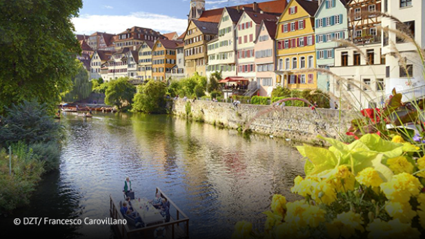 Bunte Fachwerkhäuser am Flussufer mit Stocherkahn und Blumen im Vordergrund.