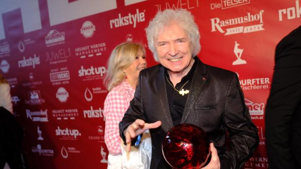 Ein Mann mit grauen Haaren hält eine rote Bowlingkugel vor einem roten Hintergrund mit Logos.