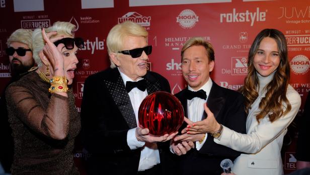 Vier elegant gekleidete Personen posieren gemeinsam und halten eine große rote Glaskugel vor einer Sponsorenwand.