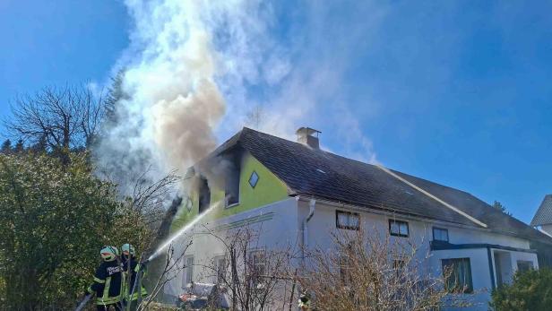 Zwei Feuerwehrmänner halten einen Schlauch und richten den Strahl auf das Fenster eines Wohnhauses, aus dem Rauch aufsteigt.