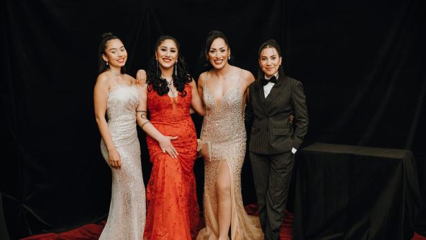 Vier elegant gekleidete Frauen posieren gemeinsam, drei in Abendkleidern und eine im Anzug, vor schwarzem Hintergrund.