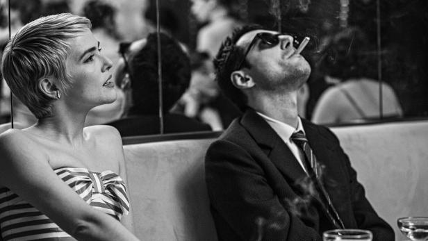 Eine Frau im gestreiften Kleid sitzt neben einem Mann im Anzug, der eine Zigarette raucht und Sonnenbrille trägt.