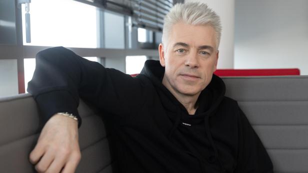 Ein Mann mit grauen Haaren und schwarzem Hoodie sitzt entspannt auf einer grauen Polsterbank vor Fenstern.