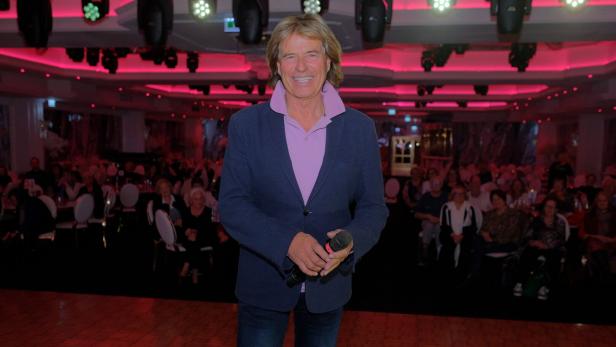 Ein Mann mit Mikrofon steht lächelnd auf einer Bühne vor einem Publikum in einem festlich beleuchteten Saal.