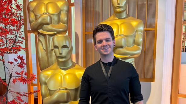 Bernhard Zimmerl steht vor drei menschengroßen Oscar-Figuren und lächelt im Koch-Outfit in die Kamera.