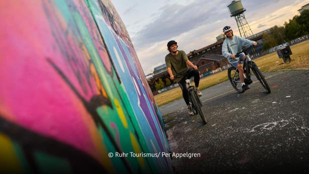 Zwei Radfahrer fahren an einer bunt besprühten Wand im urbanen Ruhrgebiet entlang.