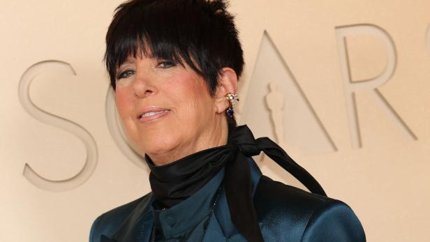 Diane Warren mit kurzem dunklem Haar, Ohrschmuck und schwarzer Schleife um den Hals vor hellem Hintergrund.