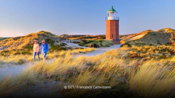 Zwei Personen spazieren bei Sonnenuntergang durch Dünenlandschaft mit kleinem Leuchtturm auf Sylt.