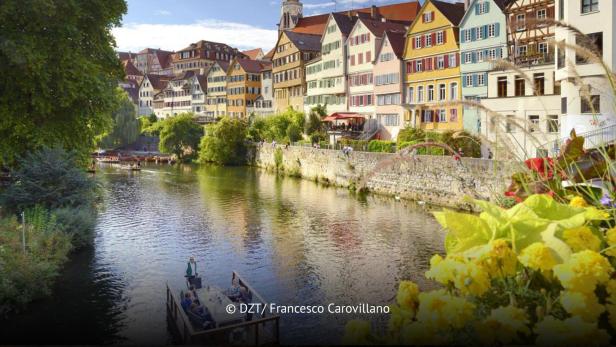 Bunte Fachwerkhäuser am Flussufer mit Stocherkahnfahrten und blühenden Blumen im Vordergrund.