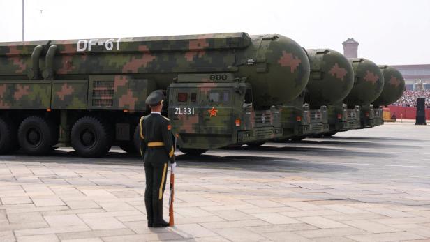 Mehrere militärische Transportfahrzeuge bei einer Militärparade fahren an einen Soldaten vorbei