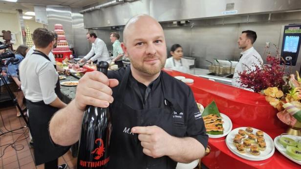 Ein Koch hält eine große schwarze Flasche mit rotem Skorpion-Logo in einer Restaurantküche, im Hintergrund arbeiten weitere Köche.