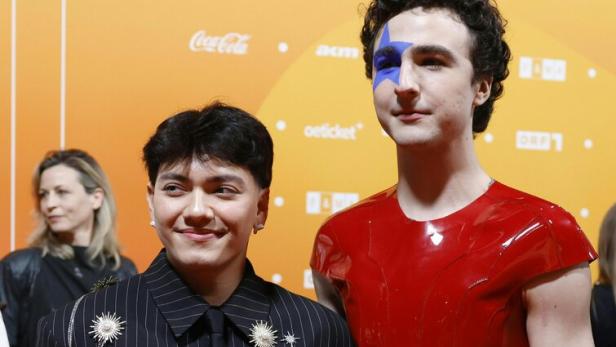 Zwei Personen posieren vor orangefarbenem Hintergrund, eine trägt einen Nadelstreifenanzug mit Schmuck, die andere ein rotes Oberteil und Gesichtsbemalung.