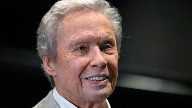 Ein Mann mit grauem Haar lächelt und trägt ein helles Jackett vor dunklem Hintergrund.