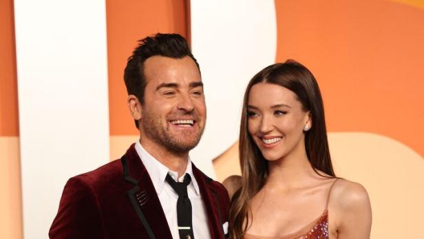 Justin Theroux im Samtjackett und Nicole Brydon Bloom im Paillettenkleid lächeln vor einem farbigen Hintergrund.