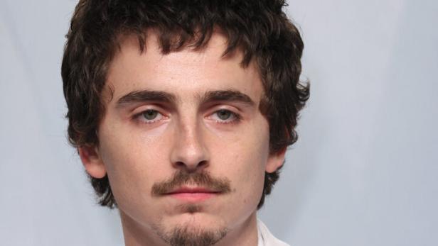 Timothée Chalamet mit dunklen, lockigen Haaren und Schnurrbart trägt einen weißen Anzug und eine auffällige Halskette.