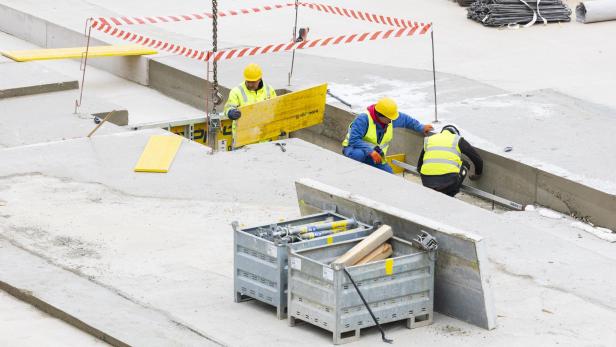 Drei Bauarbeiter mit Schutzhelmen und Warnwesten arbeiten an einer Baustelle mit Betonplatten und Baumaterialien.