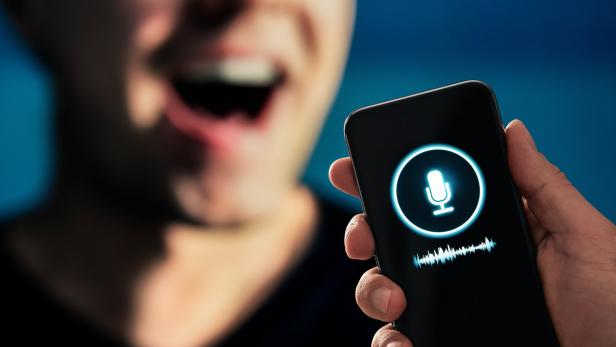 Eine Person spricht vor ein Smartphone, auf dessen Display ein Mikrofon-Symbol und eine Tonspur angezeigt werden.