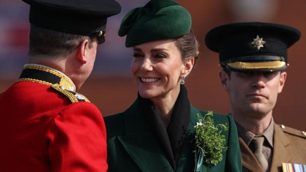 Eine elegant gekleidete Prinzessin Kate in Grün mit Hut und Anstecksträußchen spricht lächelnd mit zwei uniformierten Männern.