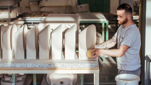 Mann mit kurz geschnittenem Haar und Tattoos schleift mit Schwamm mehrere weiße Keramikwaschbecken auf einem Tisch in einer Werkstatt