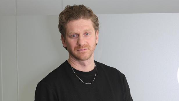 Ein Mann mit lockigen Haaren trägt ein schwarzes T-Shirt, helle Hose und zeigt mehrere Tattoos an den Armen.