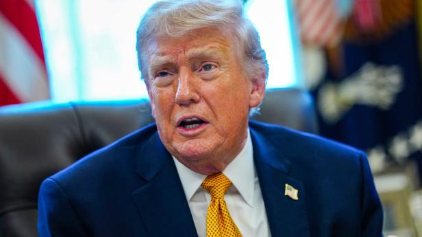 Donald Trump sitzt im Anzug und spricht, mit US-Flagge im Hintergrund.