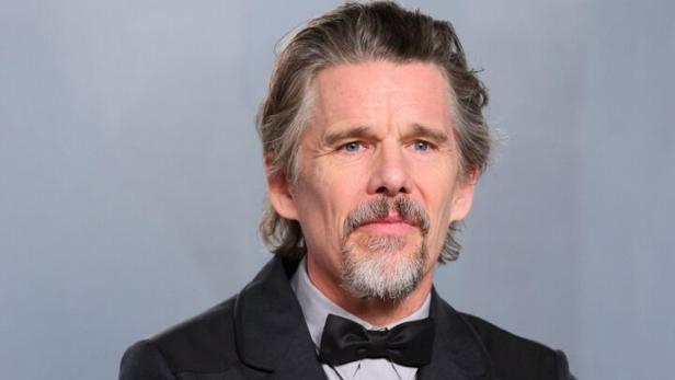 Ethan Hawke mit Fliege und Anzug verschränkt die Arme vor einem grauen Hintergrund.