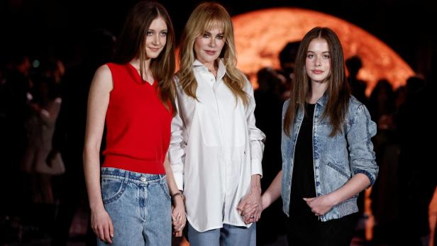 Nicole Kidman steht mit zwei jungen Frauen Händchen haltend vor einem dunklen Hintergrund.