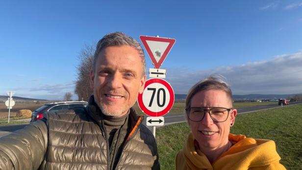 Zwei Personen stehen bei Sonnenschein neben einer Landstraße vor einem Vorfahrt-achten- und einem 70-km/h-Schild.