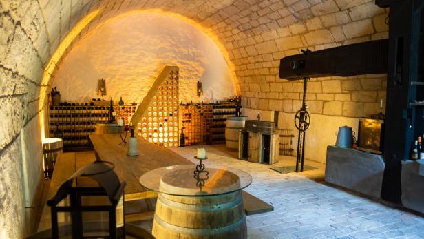 Gewölbekeller mit Weinregalen, Holztischen, Fässern und stimmungsvoller Beleuchtung.