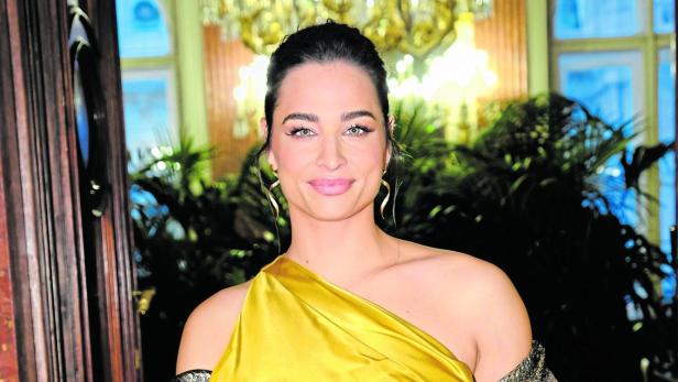 Amira Aly in goldenem, schulterfreiem Kleid mit glitzerndem Schal und Clutch.