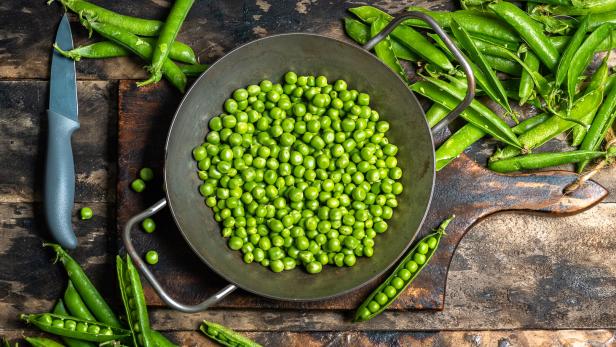 Frische grüne Erbsen liegen in einer Metallschüssel, daneben Schoten, ein Messer und ein Holzbrett auf rustikalem Untergrund.