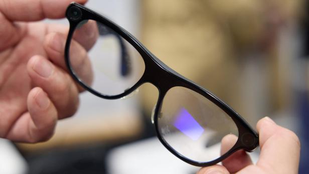 Eine schwarze Brille mit integrierten Kameras an den Ecken wird von zwei Händen gehalten.