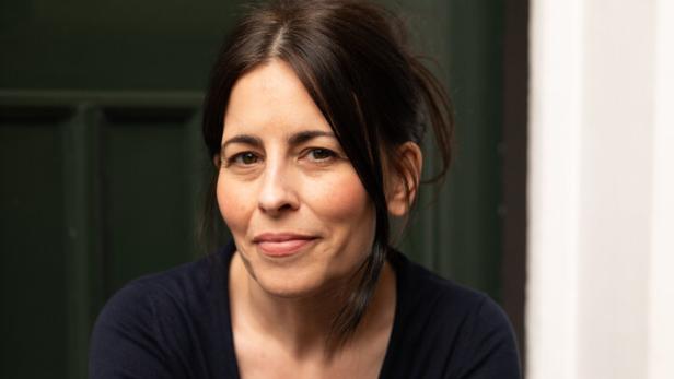 Frau mit dunklen Haaren und dunklem Pullover sitzt mit verschränkten Armen vor einer grünen Tür und lächelt leicht.