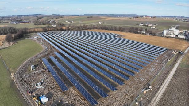 Große Solaranlage mit zahlreichen Solarpanels auf einem Feld, umgeben von ländlicher Landschaft und vereinzelten Gebäuden.
