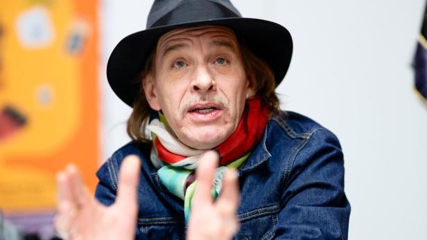 Mann mit Hut, bunter Schal und Jeansjacke gestikuliert mit den Händen und spricht, im Hintergrund unscharfe Farben.