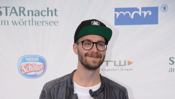 Mark Forster mit Brille, Bart, schwarzer Kappe, grauer Lederjacke und weißem T-Shirt steht vor einer Sponsorenwand.