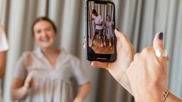 Person hält Smartphone und filmt zwei Menschen, die vor einem Vorhang posieren und tanzen.