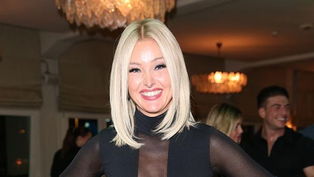 Eine lächelnde Patrice Aminati mit blondem Haar trägt einen schwarzen Rollkragenpullover und eine Hose.