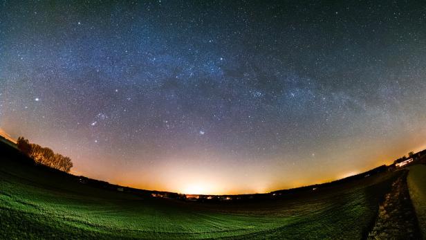 Ein weiter Sternenhimmel mit leuchtender Milchstraße über einer dunklen, leicht beleuchteten Wiesenlandschaft.